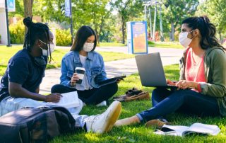 Three college students in masks studying in grass