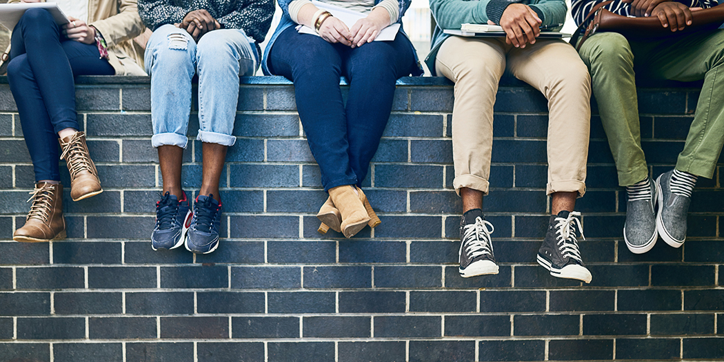 A photo of the ankles and feet of diverse students sitting on a grey brick wall