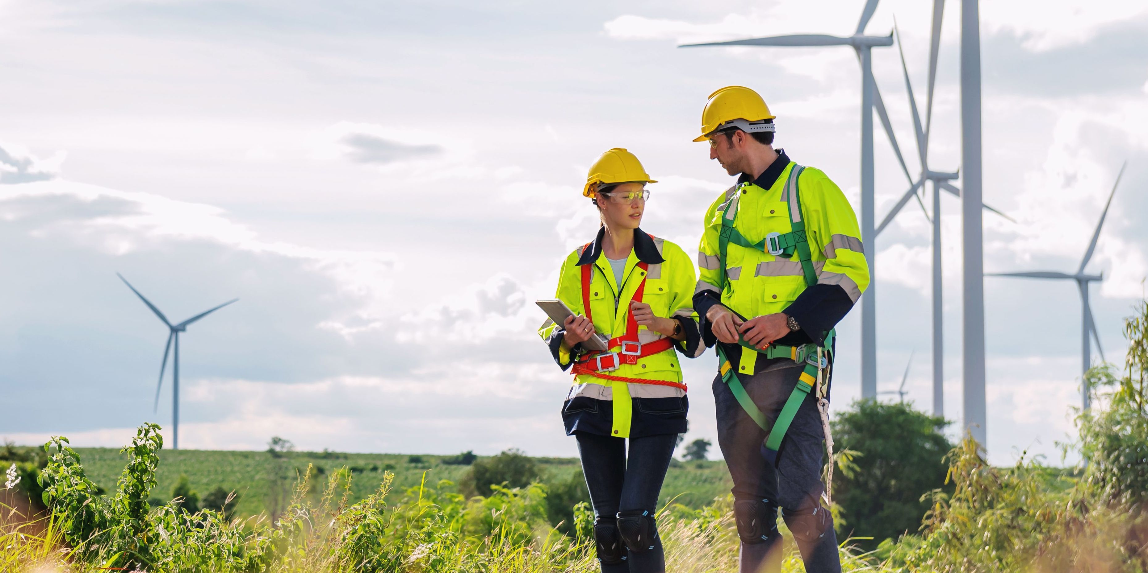 Two workers in safety gear are walking through a field of wind turbines.