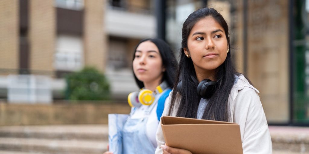 Two hispanic female students are standing outdoors on campus with books and binders