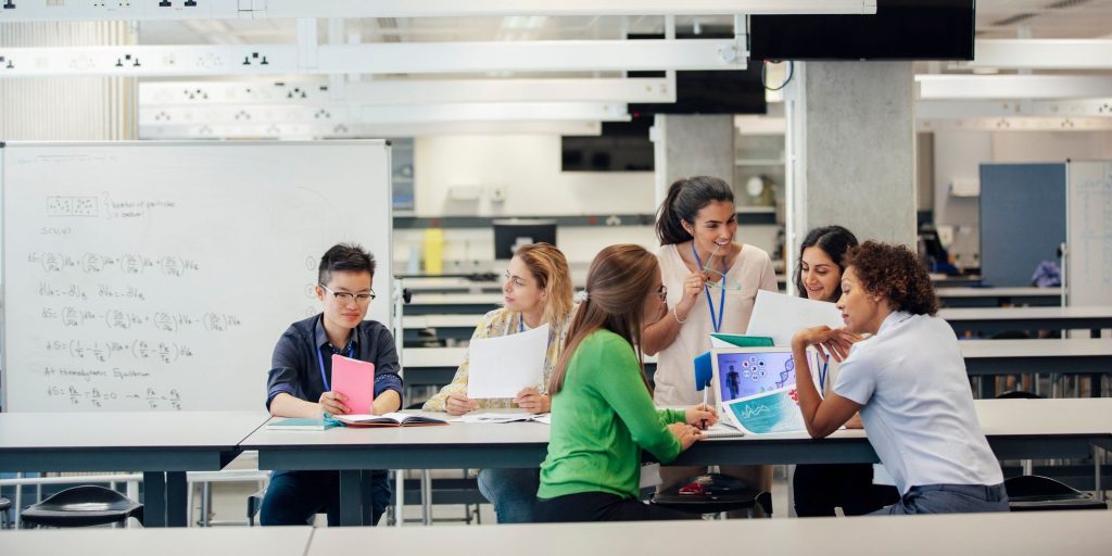 A laboratory classroom with a group of female science students working. The classmates are discussing work, and debating ideas and opinions. This architecture is well designed and versatile, allowing for changing demands in this education space. There is a whiteboard with mathematic equations on it, behind the positive animated group. The women are of differing ethnicities.