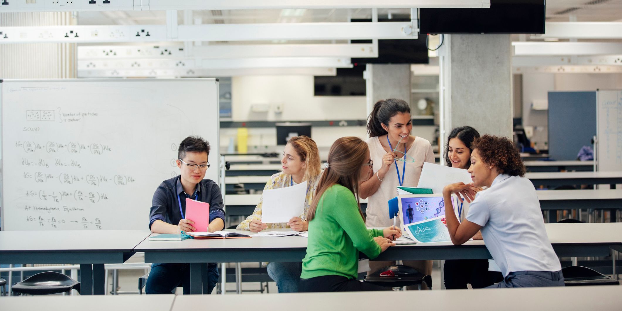 A laboratory classroom with a group of female science students working. The classmates are discussing work, and debating ideas and opinions. This architecture is well designed and versatile, allowing for changing demands in this education space. There is a whiteboard with mathematic equations on it, behind the positive animated group. The women are of differing ethnicities.